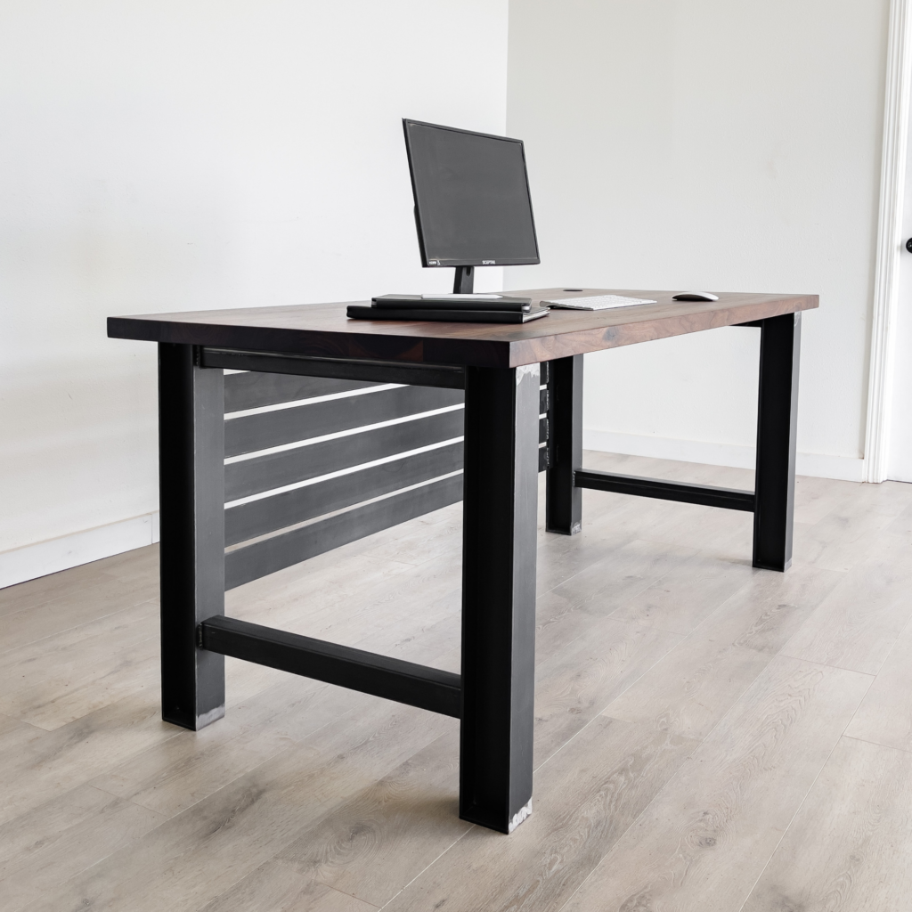 A minimalist office setup showcasing The Steele Desk, distinguished by its sleek black metal legs and providing a home for a computer monitor, keyboard, and mouse. The space is immaculately tidy and organized, set against a light neutral backdrop with wooden flooring. This piece of furniture effortlessly blends functionality with elegance.