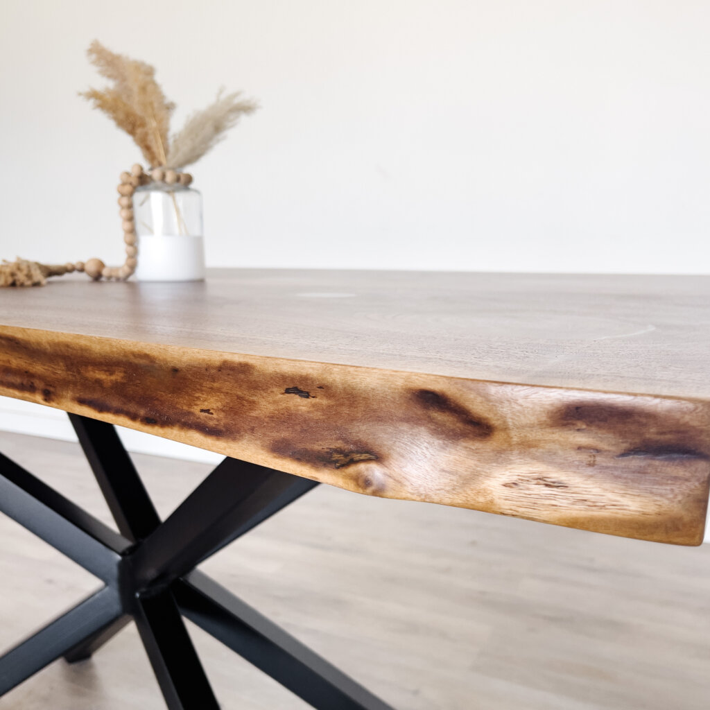 Close-up of a wooden dining table with a natural, irregular edge, showcasing the latest trends in live edge furniture. The tabletop has light wood grain patterns, and the black metal legs form an X-shape. In the background, a small white vase containing dried grasses and a string of wooden beads is placed on the table. Close-up of a wooden dining table with a natural, irregular edge, showcasing the latest trends in live edge furniture. The tabletop has light wood grain patterns, and the black metal legs form an X-shape. In the background, a small white vase containing dried grasses and a string of wooden beads is placed on the table.