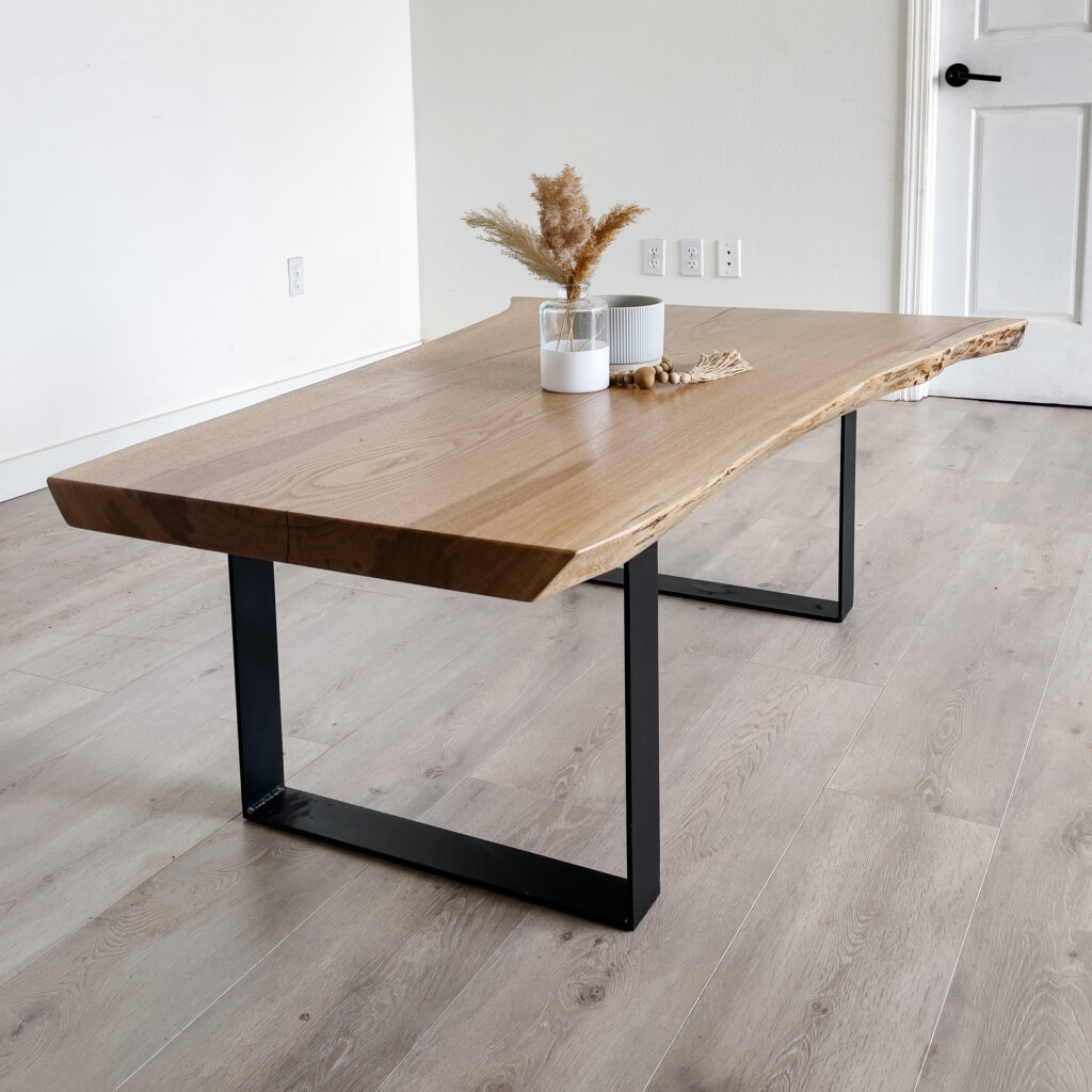 A rectangular wooden coffee table with natural live edges on the top, resting on black metal legs. On the table sits a white candle and a small beige vase containing dried pampas grass and wooden beads. The table is placed on a light wood floor in a minimalist room, embodying current trends. A rectangular wooden coffee table with natural live edges on the top, resting on black metal legs. On the table sits a white candle and a small beige vase containing dried pampas grass and wooden beads. The table is placed on a light wood floor in a minimalist room, embodying current trends.