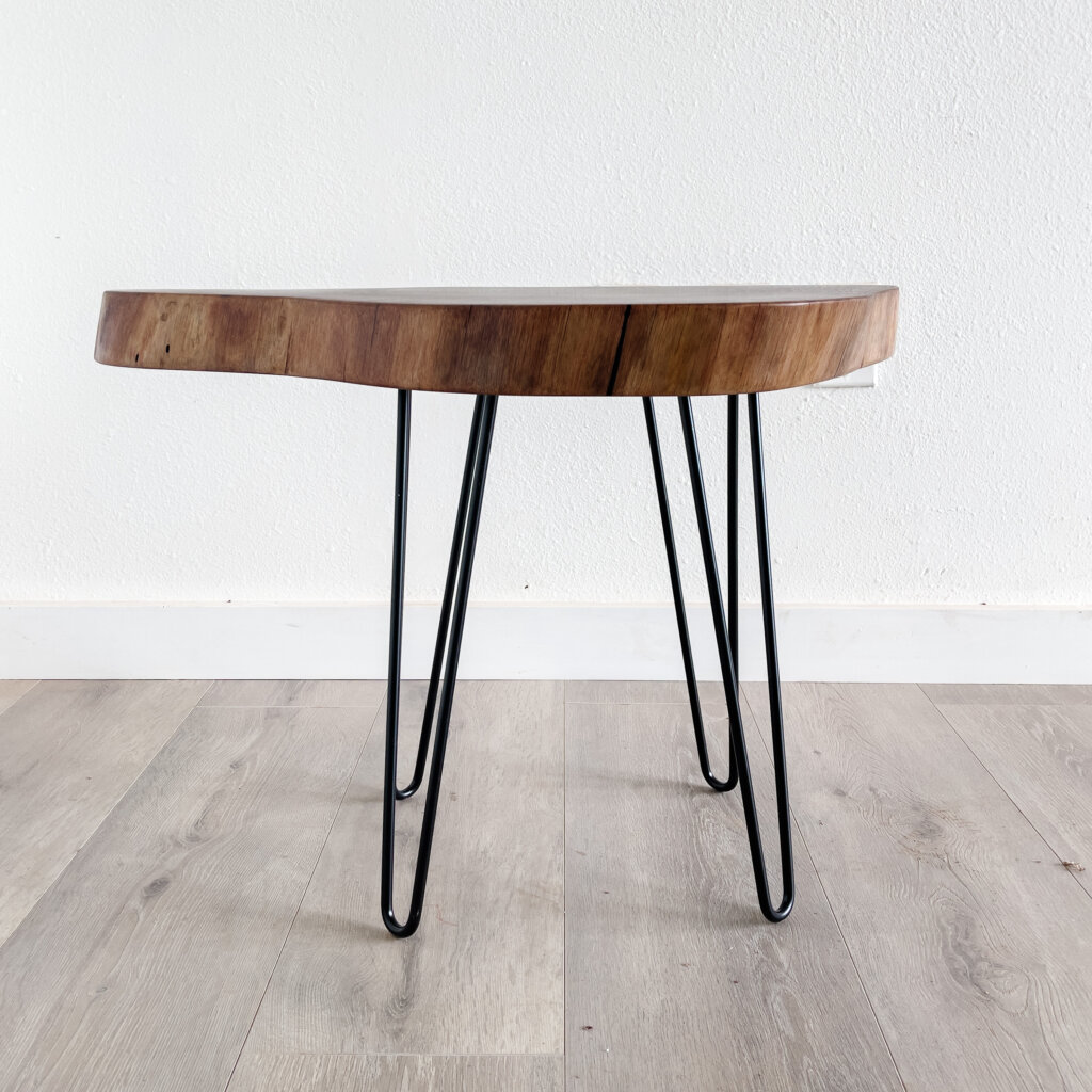 A small wooden side table with a natural, live-edge top and three black metal hairpin legs stands on a light wood floor against a plain white wall. This piece of Live Edge Furniture perfectly captures minimalist and modern trends. A small wooden side table with a natural, live-edge top and three black metal hairpin legs stands on a light wood floor against a plain white wall. This piece of Live Edge Furniture perfectly captures minimalist and modern trends.