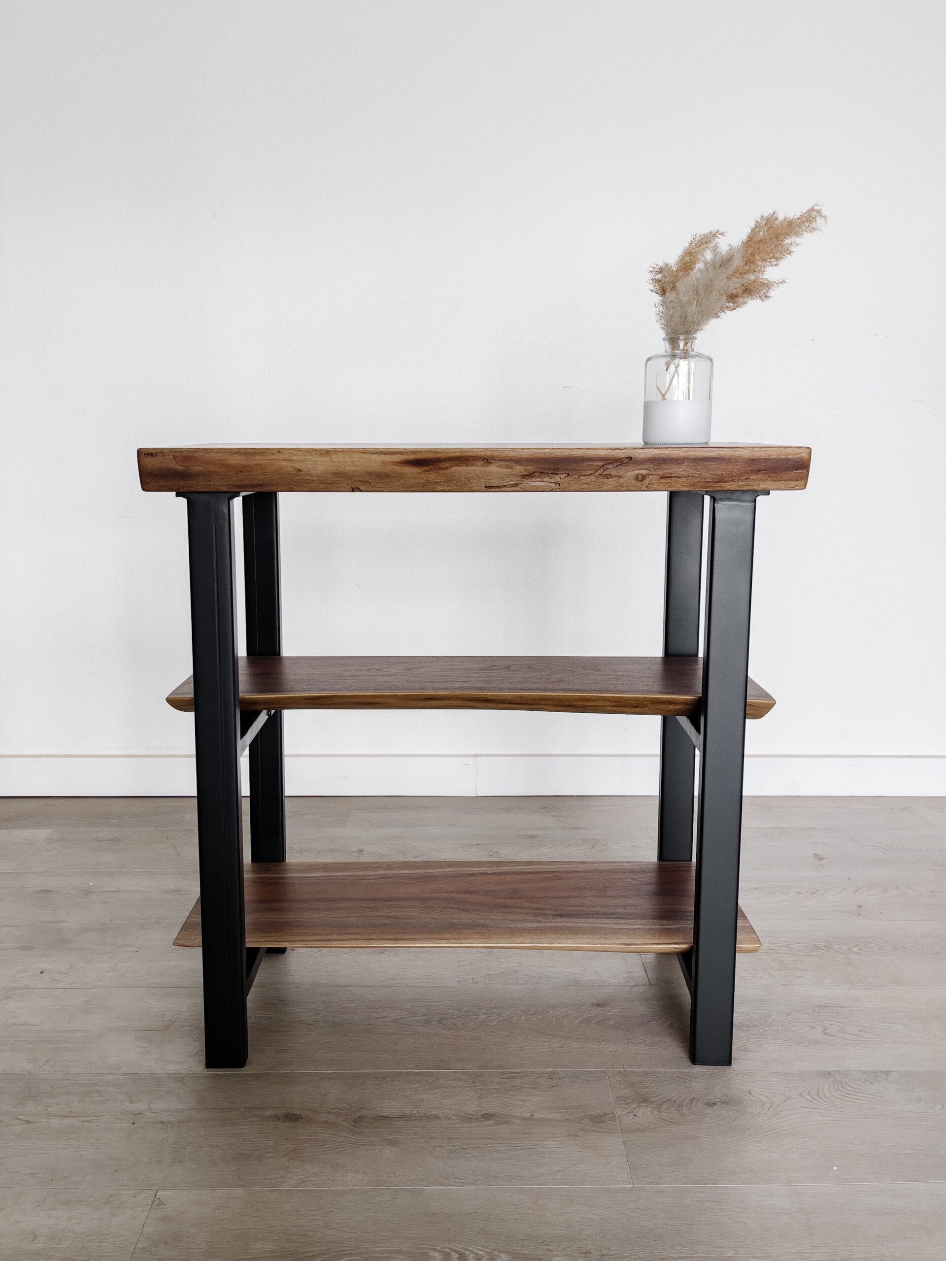 A three-tiered wooden shelf with black metal legs stands against a plain white wall on a light wood floor. A glass vase with dried pampas grass decorates the top shelf, adding a minimalist touch to the modern furniture piece, perfectly aligning with current live edge furniture trends. A three-tiered wooden shelf with black metal legs stands against a plain white wall on a light wood floor. A glass vase with dried pampas grass decorates the top shelf, adding a minimalist touch to the modern furniture piece, perfectly aligning with current live edge furniture trends.