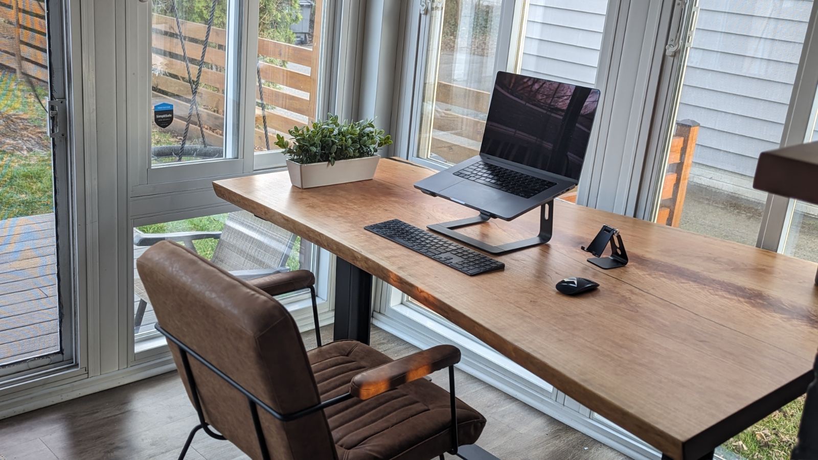 A neatly arranged home office setup by a large window features the latest in furniture trends. The desk holds a laptop on a stand, a keyboard, a mouse, and a small potted plant, all atop an elegant piece of live edge furniture. A brown cushioned chair is placed in front of the desk. Natural light floods the room from the surrounding windows. A neatly arranged home office setup by a large window features the latest in furniture trends. The desk holds a laptop on a stand, a keyboard, a mouse, and a small potted plant, all atop an elegant piece of live edge furniture. A brown cushioned chair is placed in front of the desk. Natural light floods the room from the surrounding windows.