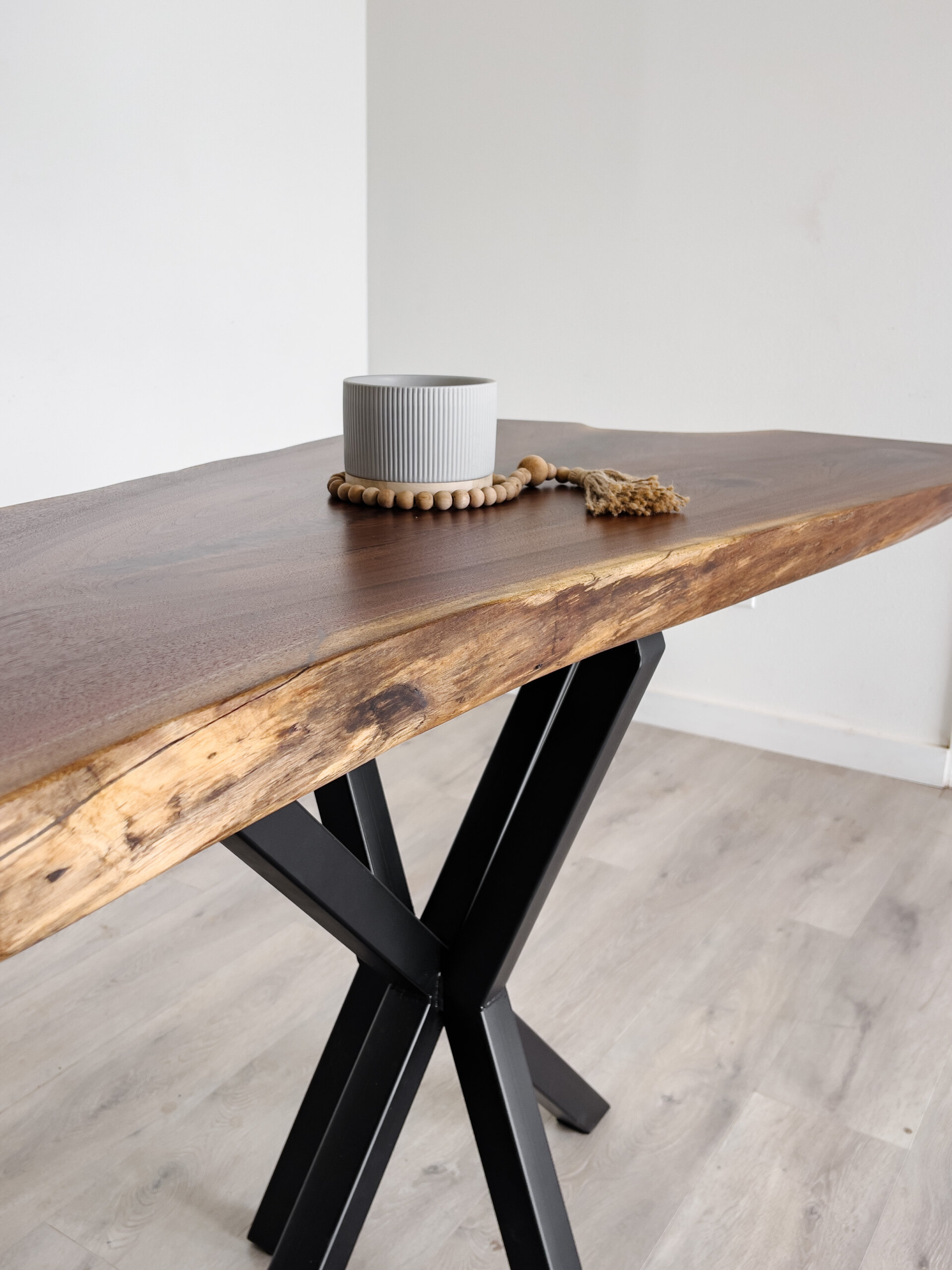 A modern wooden table with a live edge, held by black metal legs, stands on a light wood floor. A small gray ceramic pot and a wooden bead garland with a tassel are placed atop the table. Embracing current furniture trends, this piece shines against the plain white wall in the background. A modern wooden table with a live edge, held by black metal legs, stands on a light wood floor. A small gray ceramic pot and a wooden bead garland with a tassel are placed atop the table. Embracing current furniture trends, this piece shines against the plain white wall in the background.