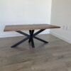 The "Ready To Ship - Black Walnut Coffee Table," featuring a rectangular design with a thick tabletop and black metal legs that form a geometric pattern, is situated against a plain white wall in a corner of a room with light-colored wooden flooring. Electrical outlets are visible on the wall to the right.