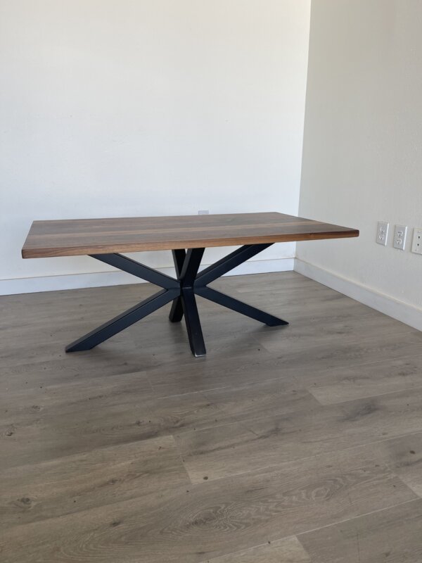 The "Ready To Ship - Black Walnut Coffee Table," featuring a rectangular design with a thick tabletop and black metal legs that form a geometric pattern, is situated against a plain white wall in a corner of a room with light-colored wooden flooring. Electrical outlets are visible on the wall to the right.