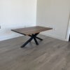 A "Ready To Ship - Black Walnut Coffee Table" with a modern minimalist design features a rectangular black walnut top and black X-shaped legs. It sits elegantly on a gray wooden floor against a plain white wall, enhancing the clean and uncluttered space around it, and is conveniently available for immediate shipping.