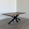 A wooden dining table with a dark finish and a modern design featuring an X-shaped metal base stands against a light-colored wall on a hardwood floor, complementing the Ready To Ship - Black Walnut Coffee Table.