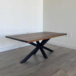 A wooden dining table with a dark finish and a modern design featuring an X-shaped metal base stands against a light-colored wall on a hardwood floor, complementing the Ready To Ship - Black Walnut Coffee Table.