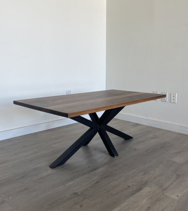 A wooden dining table with a dark finish and a modern design featuring an X-shaped metal base stands against a light-colored wall on a hardwood floor, complementing the Ready To Ship - Black Walnut Coffee Table.