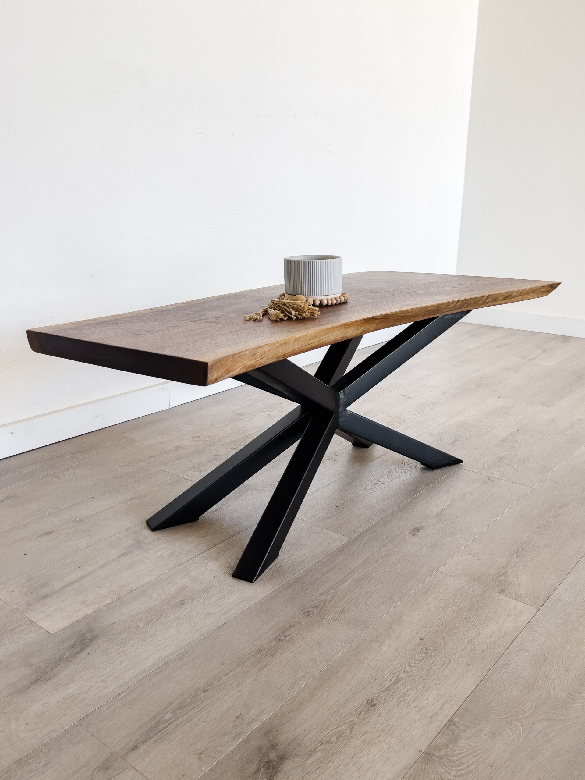 A stunning piece of live edge furniture, this wooden table boasts a natural edge top and a black, geometric X-shaped metal base. Positioned on a light-colored wooden floor, it showcases a gray ribbed cylindrical candle, a beaded decoration, and a small bundle of dried wheat. A stunning piece of live edge furniture, this wooden table boasts a natural edge top and a black, geometric X-shaped metal base. Positioned on a light-colored wooden floor, it showcases a gray ribbed cylindrical candle, a beaded decoration, and a small bundle of dried wheat.