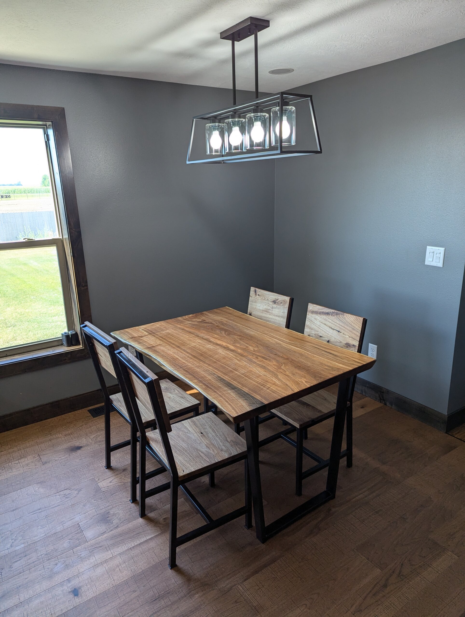 A modern dining room with a rectangular live edge wooden table and four matching chairs. The room has gray walls, a wooden floor, a window with a view of green grass outside, and an elegant rectangular light fixture hanging from the ceiling. A modern dining room with a rectangular live edge wooden table and four matching chairs. The room has gray walls, a wooden floor, a window with a view of green grass outside, and an elegant rectangular light fixture hanging from the ceiling.