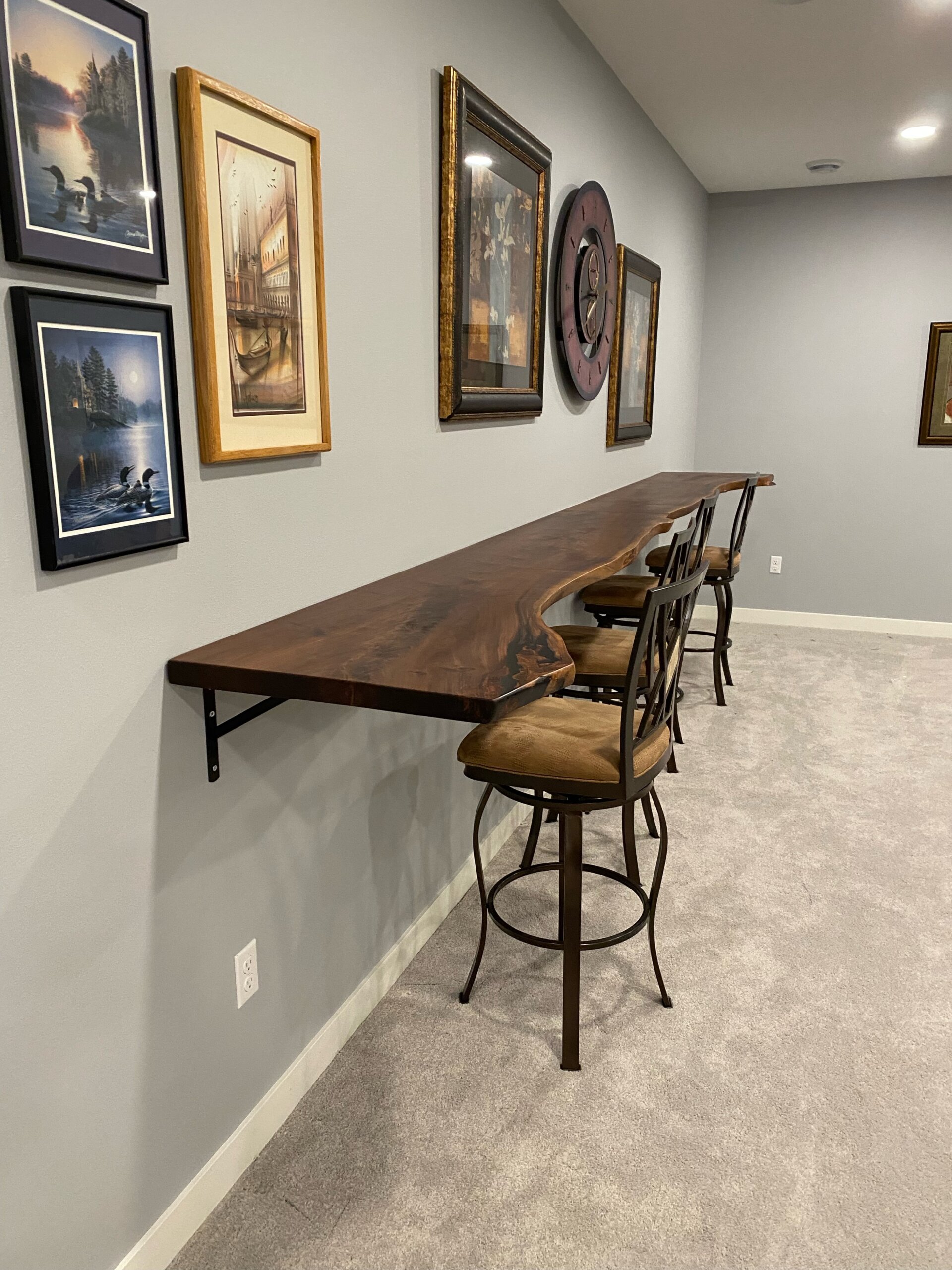 A minimalist room features a high wooden counter with a live edge, wall-mounted with black brackets. Three brown cushioned bar stools with ornate metal backs are placed along the counter. Reflecting the latest furniture trends, the walls are adorned with framed artwork and paintings. A minimalist room features a high wooden counter with a live edge, wall-mounted with black brackets. Three brown cushioned bar stools with ornate metal backs are placed along the counter. Reflecting the latest furniture trends, the walls are adorned with framed artwork and paintings.