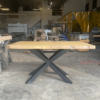 A Ready To Ship - Rainbow Poplar Single Slab Dakota Table, featuring a rectangular wooden top and metal X-shaped legs, stands in the center of a workshop. Various pieces of lumber, tools, and machinery are visible in the background, along with a partially transparent yellow curtain. The floor is dusty. This ready-to-ship piece awaits its new owner.