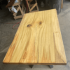 The Ready To Ship - Rainbow Poplar Single Slab Dakota Table features a rectangular shape with a light, natural finish that highlights the prominent wood grain, knots, and cracks. Supported by metal legs, this distinctive piece is situated in a workshop surrounded by various tools and materials.