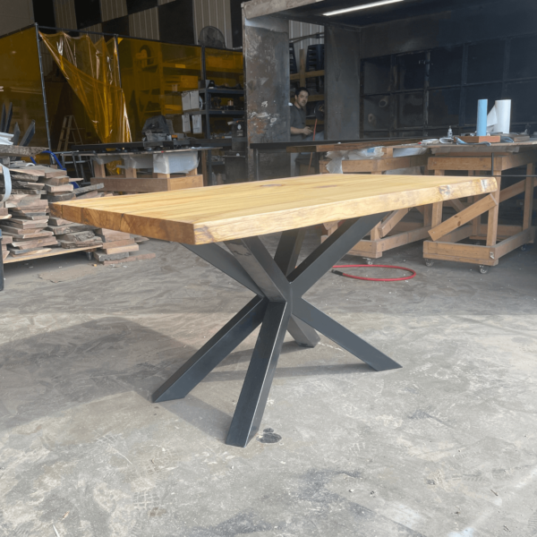 A "Ready To Ship - Rainbow Poplar Single Slab Dakota Table" with a natural finish sits on a floor in a workshop. This wooden table boasts a thick rectangular top crafted from a single slab of Rainbow Poplar and features a black metal base with intersecting legs. Various woodworking tools, materials, and machinery are visible in the background.