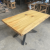 The Ready To Ship - Rainbow Poplar Single Slab Dakota Table, featuring a natural, unfinished surface and a light tan color with visible wood grain and knots, stands on a concrete floor in a workshop. Its tabletop is supported by X-shaped black metal legs, while other woodworking tools and materials are visible in the background.
