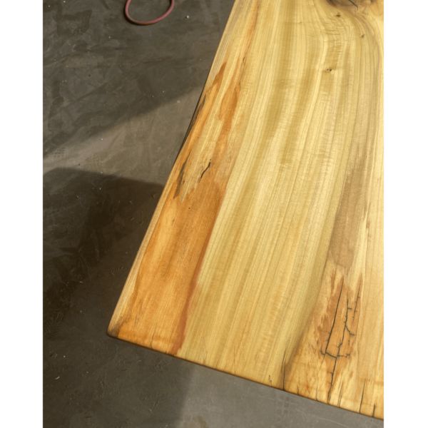 A close-up view of the Ready To Ship - Rainbow Poplar Single Slab Dakota Table surface crafted from a single slab of Rainbow Poplar, showcasing its natural, polished finish. The wood displays various grain patterns and color variations ranging from light to dark tones. The table edge is rounded, set against a dark, textured floor background.
