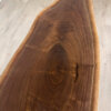 A close-up shot of the "Ready To Ship - Black Walnut Live Edge Coffee Table" reveals its polished black walnut surface with natural wood grain patterns. The live edge table retains an organic shape, showcasing rich, dark hues and a smooth finish with visible rings and textures, displayed on a light-colored floor.