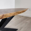 A close-up of the Ready To Ship - Black Walnut Live Edge Coffee Table highlights its natural, uneven edge. The table rests on angled black metal legs, with a light, glossy finish on the surface. The background features a white wall and light-colored wooden floor.