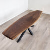 The Ready To Ship - Black Walnut Live Edge Coffee Table, showcasing a rich dark brown surface and visible wood grain patterns, stands on a light wood floor. This elegant table is supported by a black metal base with crisscrossing legs. The background features a plain white wall.