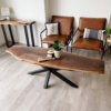 A modern living room showcases a stunning Ready To Ship - Black Walnut Live Edge Coffee Table with a sleek metal base. On the table sits a small potted plant and a candle. Behind the table are two brown leather chairs with decorative pillows. In the corner, there's a matching console table.