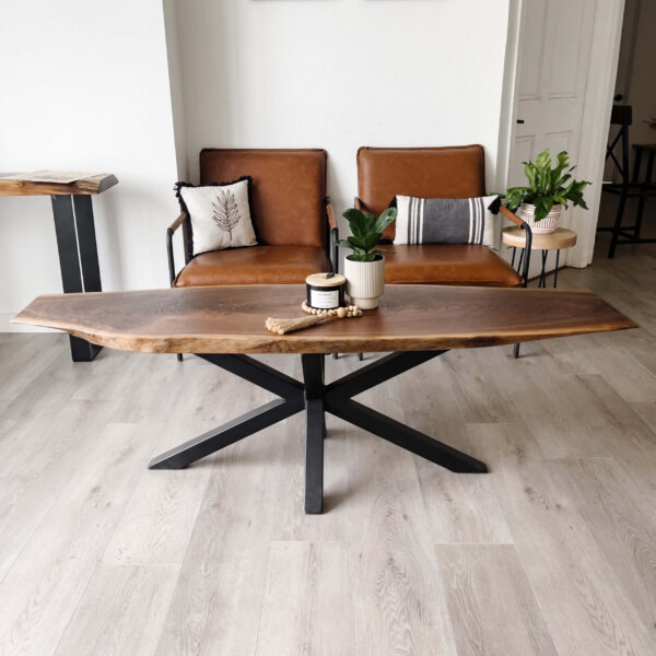 A modern living room showcases the Ready To Ship - Black Walnut Live Edge Coffee Table featuring black metal legs. Behind it, two brown leather chairs with cushions are perfectly placed. The table is accessorized with two small potted plants and various decorative items. The room itself boasts light-colored flooring and minimalistic decor.