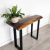 A striking Black Walnut Live Edge Coffee Table from our Ready To Ship collection stands against a white wall. It features black metal legs and a polished wood surface that highlights its rustic charm. Adding to its allure, a potted plant with broad green leaves in a stylish white and pink striped pot adorns the table. The scene is completed by a light wood floor.
