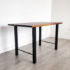 The Ready To Ship - Black Walnut Desk With H-Shaped Legs, featuring a minimalist rectangular top and sleek black metal legs, is positioned on a light-colored wooden floor against a plain white wall in a sparsely furnished room. It seamlessly blends into any modern space.