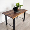 A minimalist workspace setup features the "Ready To Ship - Black Walnut Desk With H-Shaped Legs," showcasing a sleek laptop open on the tabletop next to a potted green plant. The room is adorned with light-colored wooden flooring and a white wall background, creating a clean and modern ambiance. Ready to ship today!