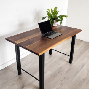 A minimalist workspace setup features the "Ready To Ship - Black Walnut Desk With H-Shaped Legs," showcasing a sleek laptop open on the tabletop next to a potted green plant. The room is adorned with light-colored wooden flooring and a white wall background, creating a clean and modern ambiance. Ready to ship today!