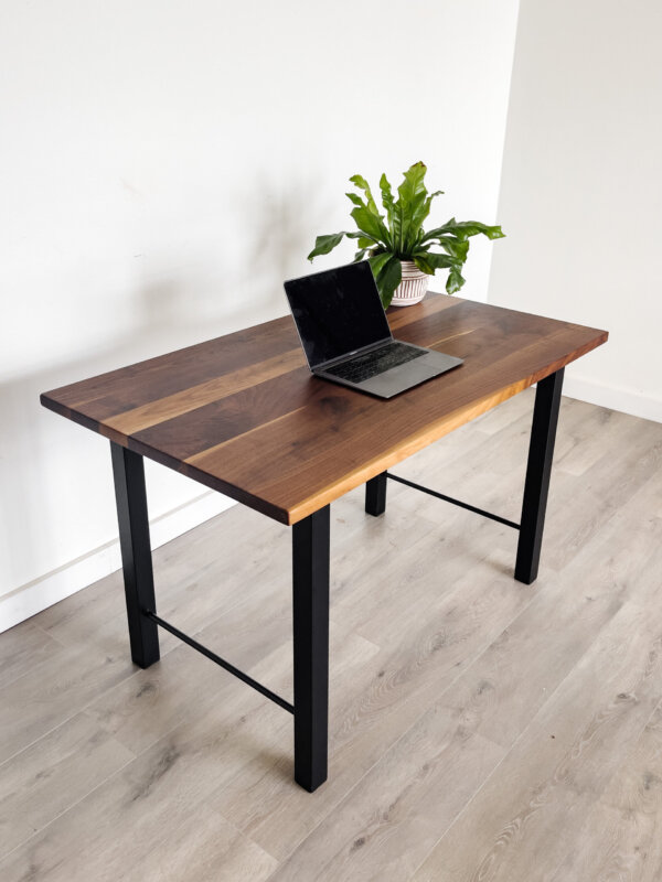 A minimalist workspace setup features the "Ready To Ship - Black Walnut Desk With H-Shaped Legs," showcasing a sleek laptop open on the tabletop next to a potted green plant. The room is adorned with light-colored wooden flooring and a white wall background, creating a clean and modern ambiance. Ready to ship today!