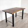 A Ready To Ship - Black Walnut Desk With H-Shaped Legs stands on light wood flooring against a plain white wall. The desk, featuring a dark brown top made of black walnut and sturdy H-shaped black metal legs, showcases a rectangular shape with a simple, modern design.