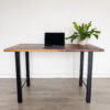 Against a white wall on a wooden floor stands the Ready To Ship Black Walnut Desk with H-shaped black metal legs. Centered on the desk is a closed laptop, and to its right sits a potted green plant, creating an overall setting that is clean and simple.