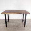 The Ready to Ship - Black Walnut Desk With H-Shaped Legs features a polished, multi-toned surface and four sleek H-shaped legs. Positioned against a plain white wall on a light gray wooden floor, this desk is ready to ship for your convenience.