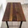 A Ready To Ship - Black Walnut Desk with a polished surface featuring contrasting grain patterns stands on sleek black, H-shaped legs. The desk sits on a light wood floor in a minimalist room with white walls, a door, and electrical outlets in the background.
