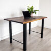 A Ready To Ship - Black Walnut Desk With H-Shaped Legs stands on a light-colored hardwood floor. The modern desk, featuring sleek black metal legs, holds a laptop and a potted green plant against a plain white wall.