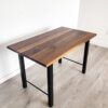 Introducing the Ready To Ship - Black Walnut Desk With H-Shaped Legs: a sleek rectangular piece boasting a sophisticated two-tone brown finish on its tabletop. The desk features H-shaped black metal legs and includes a support bar between the legs on the shorter sides, all set against a light wood floor by a plain white wall to create an effortlessly modern vibe.