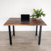 A minimalist setup featuring the "Ready To Ship - Black Walnut Desk With H-Shaped Legs," a closed laptop, and a potted green plant on the right side of the desk. The background includes a white wall and light-colored wooden flooring, making this elegant space ready for use.