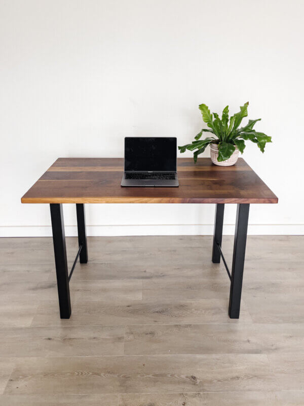 A minimalist setup featuring the "Ready To Ship - Black Walnut Desk With H-Shaped Legs," a closed laptop, and a potted green plant on the right side of the desk. The background includes a white wall and light-colored wooden flooring, making this elegant space ready for use.