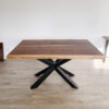 The "Ready To Ship - Book Matched Black Walnut Dakota Table" features a modern design with a rectangular black walnut top and an X-shaped black metal base. Set on a light-colored wooden floor, this table boasts a minimalistic and clean background, making it perfect for any decor. Ready to ship for your convenience.
