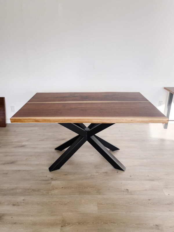 The "Ready To Ship - Book Matched Black Walnut Dakota Table" features a modern design with a rectangular black walnut top and an X-shaped black metal base. Set on a light-colored wooden floor, this table boasts a minimalistic and clean background, making it perfect for any decor. Ready to ship for your convenience.