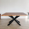 The Ready To Ship - Book Matched Black Walnut Dakota Table features a wooden dining surface with a rectangular top and a distinctive X-shaped black steel base. Crafted from black walnut, it is positioned on a light wood floor against a plain white wall, offering a modern and minimalist aesthetic.
