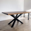 A rectangular Black Walnut dining table with a natural finish and live edge sits on a floor with light wood planks. The Ready To Ship - Book Matched Black Walnut Dakota Table features a black, metal X-shaped base. A portion of another table or a bench can be seen in the background near the wall, and it is ready to ship.