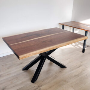 A Ready To Ship - Book Matched Black Walnut Dakota Table, featuring a dark brown, polished Black Walnut surface and black metal X-shaped legs, is showcased on a light wood floor. Another similar table can be partially seen in the background against a white wall. This elegant piece is poised to elevate any space with its sleek design.