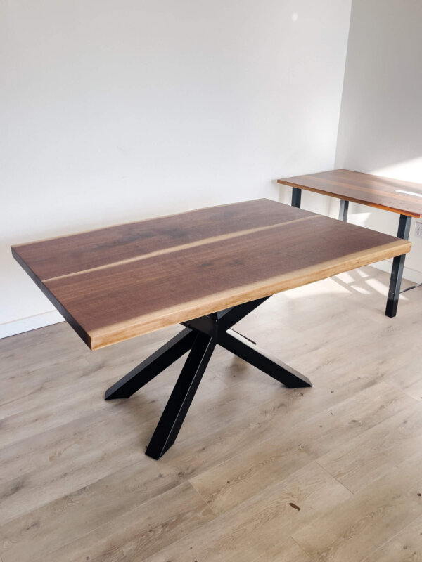 A Ready To Ship - Book Matched Black Walnut Dakota Table, featuring a dark brown, polished Black Walnut surface and black metal X-shaped legs, is showcased on a light wood floor. Another similar table can be partially seen in the background against a white wall. This elegant piece is poised to elevate any space with its sleek design.