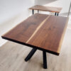 A Ready To Ship - Book Matched Black Walnut Dakota Table, featuring a natural, smooth surface with dark brown grain and black metal legs forming an X shape, sits on light wood plank flooring. Behind it, a smaller wooden desk with similar legs rests near a white wall.