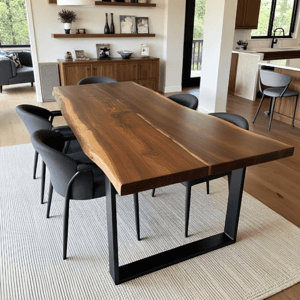 A modern dining area with a large, natural wood table and black metal legs. Surrounded by six gray upholstered chairs, the setup rests on a light-colored rug. The background shows a wooden sideboard, decor items, and a kitchen area.
