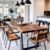 A modern dining room features a long wooden table with black metal legs and eight brown chairs. Overhead, three black pendant lights hang. The room includes a view of a living area, large windows, and a kitchen island with stools in the background.
