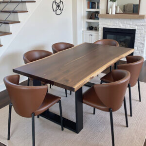 Dining room with a modern wooden table surrounded by six brown leather chairs on a light-colored rug. A staircase with wooden steps and metal railing is visible to the left, and a fireplace with a mirror and bookshelf is on the right.