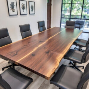 A conference room with a large wooden table surrounded by eight black leather chairs. Three framed botanical prints hang on the white wall. A large window offers a view of trees outside, letting in natural light. The floor is light wood.